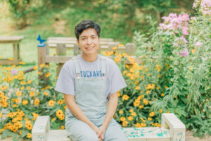 Man wearing Tuckahoe shirt and overalls, sitting in front of yellow flowers.